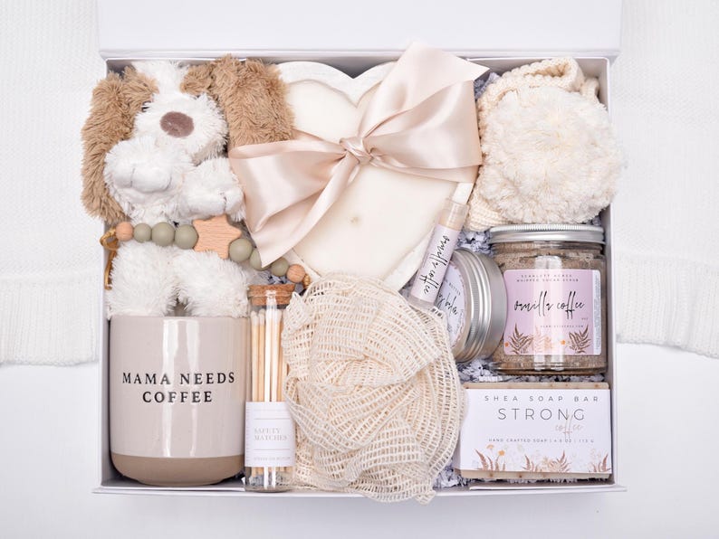 May include: A gift box containing a plush dog toy, a heart-shaped candle with a satin ribbon, a mug labeled MAMA NEEDS COFFEE, a glass tube of matches, a mesh sponge, a lip balm, a jar of vanilla coffee scrub, and a bar of shea soap labeled STRONG COFFEE.