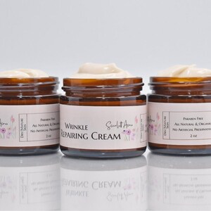 May include: Three amber glass jars of face cream with white labels. The middle jar says "Wrinkle Repairing Cream" and the other two jars say "Cream". All jars have a floral design and the text "Scarlet Acne" on the label. The jars are on a white surface.