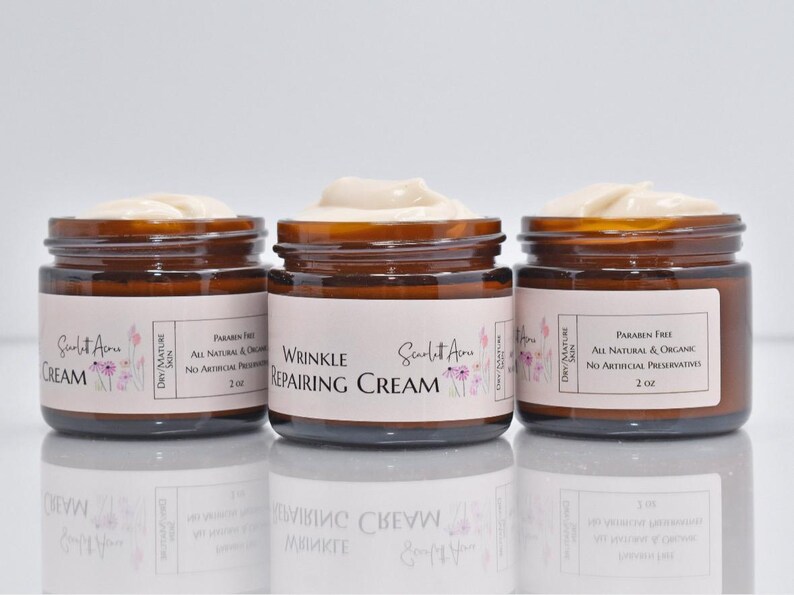 May include: Three amber glass jars of face cream with white labels. The middle jar says "Wrinkle Repairing Cream" and the other two jars say "Cream". All jars have a floral design and the text "Scarlet Acne" on the label. The jars are on a white surface.