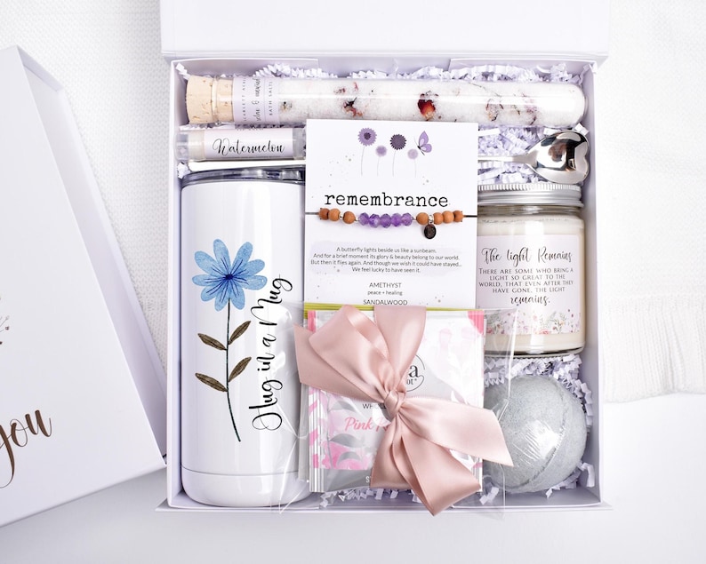 Puede incluir: Una caja de regalo blanca llena de art&iacute;culos de cuidado personal. Incluye un vaso con una flor azul y el texto "Hug in a Mug", una vela, una bomba de ba&ntilde;o, una pulsera, sales de ba&ntilde;o y un b&aacute;lsamo labial.