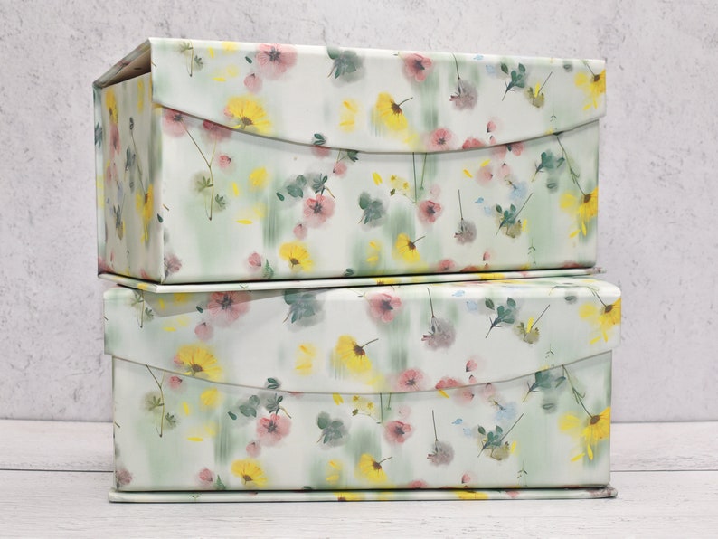 May include: Two stackable storage boxes with a floral pattern. The boxes are white with a green, yellow, and pink floral design.