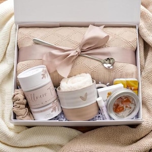 May include: A white gift box containing a beige knit blanket tied with a satin ribbon and a silver heart-shaped spoon. Included are a Wellness Tea canister, a two-toned ceramic mug with a heart, a lip balm, and a small tin of orange slices.