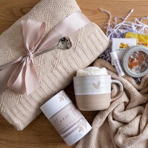 May include: A gift set featuring a beige knit blanket tied with a satin ribbon, a heart-shaped spoon, a two-toned ceramic mug with a heart icon, a Wellness Tea canister, a lip balm, and a small round tin containing orange-colored candies.