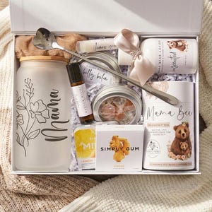 May include: A white gift box containing a frosted glass tumbler with a bamboo lid and floral Mama text, a silver heart-shaped spoon, a roller bottle, a belly balm tin, a lotion bottle with a pink ribbon, pregnancy tea, and Simply Gum ginger mints.