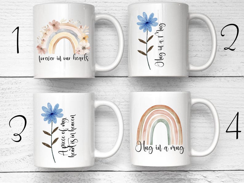 Puede incluir: Cuatro tazas de cer&aacute;mica blanca con diferentes dise&ntilde;os. Una taza presenta un arco&iacute;ris y un dise&ntilde;o floral con el texto "forever in our hearts". Otra tiene una flor azul y el texto "Hug in a Mug". La tercera taza tiene una flor azul y el texto "A piece of my heart is in heaven". La &uacute;ltima taza tiene un arco&iacute;ris y el texto "Hug in a mug."