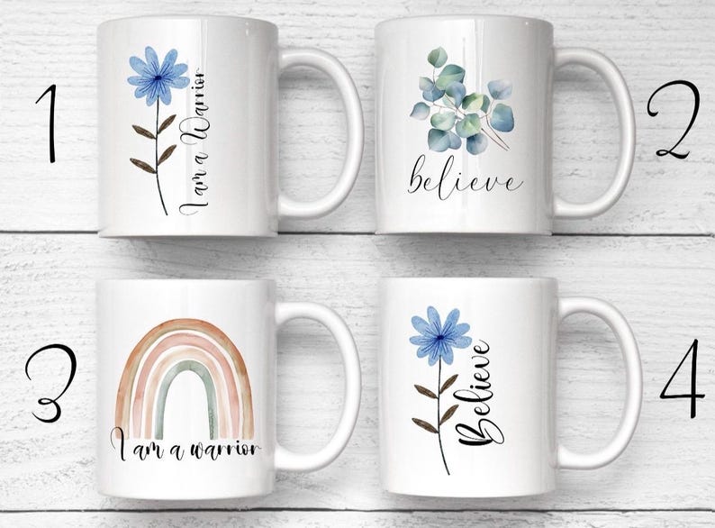 Op de afbeelding: Vier witte keramische mokken gerangschikt in een raster. Mok 1 heeft een blauwe bloem en de tekst 'I am a Warrior'. Mok 2 toont groene eucalyptusbladeren en het woord 'believe'. Mok 3 toont een gedempte regenboog en 'I am a warrior'. Mok 4 heeft een blauwe bloem en 'Believe'.