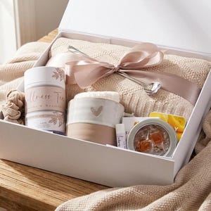 Op de afbeelding: Een witte geschenkdoos met een beige gebreide deken, een cilindervormige houder met het label Wellness Tea, een tweekleurige keramische mok met een hartje, een zilveren lepel, een lippenbalsem, een rond blikje en een haarelastiekje.