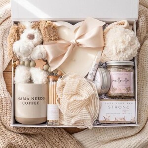 May include: A gift box containing a plush dog, a heart-shaped item with a beige ribbon, a beige mug labeled MAMA NEEDS COFFEE, a glass vial of matches, a mesh sponge, a lip balm, a small tin, a jar of vanilla coffee scrub, and a shea soap bar.