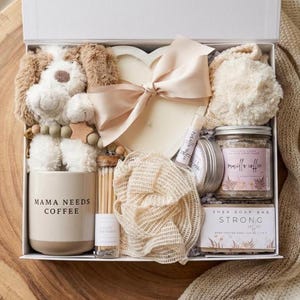 May include: A gift box containing a plush dog, a heart-shaped candle with a beige ribbon, a beige mug labeled MAMA NEEDS COFFEE, a glass vial of matches, a mesh sponge, a lip balm, a jar of vanilla coffee scrub, and a bar of strong coffee soap.