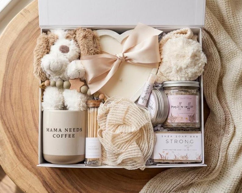 May include: A gift box containing a plush dog, a heart-shaped candle with a beige ribbon, a beige mug labeled MAMA NEEDS COFFEE, a glass vial of matches, a mesh sponge, a lip balm, a jar of vanilla coffee scrub, and a bar of strong coffee soap.
