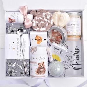 May include: A white gift box filled with self-care items. Includes a jar of citrus coconut scrub, a Mama Bear tea tin, a Mama water bottle, an eye pillow, and other items. A pink ribbon and a spoon are also included.