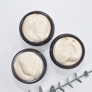 May include: Three small jars of white cream with brown lids. The jars are on a white surface with a sprig of green leaves.