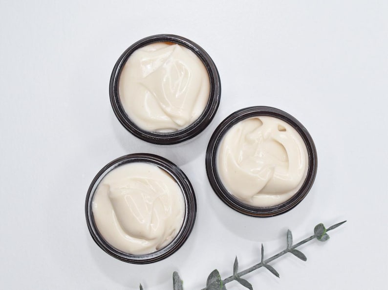 May include: Three small jars of white cream with brown lids. The jars are on a white surface with a sprig of green leaves.