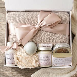 Op de afbeelding: Een witte geschenkdoos met een beige gebreide deken vastgebonden met een satijnen lint, een fles hand- en bodylotion, een ronde badbom, een net-spons, een kaars met het label You are Starry, lippenbalsem en een stuk eucalyptus en munt sheazeep.
