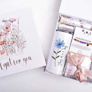 Puede incluir: Una caja de regalo blanca con un dise&ntilde;o floral y el texto "A gift for you". En el interior, un vaso blanco con una flor azul y las palabras "Hug in a Mug", una pulsera, una vela y otros art&iacute;culos.