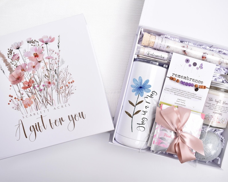 Puede incluir: Una caja de regalo blanca con un dise&ntilde;o floral y el texto "A gift for you". En el interior, un vaso blanco con una flor azul y las palabras "Hug in a Mug", una pulsera, una vela y otros art&iacute;culos.