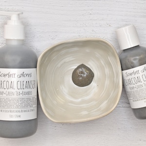 Op de afbeelding: Twee flessen met houtskoolreiniger, een met een pompdispenser en een met een schroefdop, beide met het label "Scarlett Acres Charcoal Cleanser Hemp Green Tea Bamboo". Een kleine witte kom met een scheutje houtskoolreiniger erin.