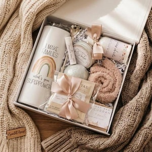 May include: A white gift box containing a tall tumbler with a rainbow graphic and the text I MAKE FAMILIES what's your SUPERPOWER, a lip balm, a round candle tin, a bath bomb, a beige fuzzy item, a tea sachet with a pink ribbon, and a soap bar.