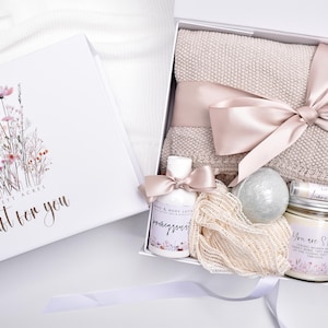 Op de afbeelding: Een witte geschenkdoos met een bloemenillustratie en de tekst Scarlett Acres A gift for you. Binnenin zit een beige gebreide deken met een satijnen lint, een flesje lotion, een bruisbal, lippenbalsem, een kaars en een netvormige spons.