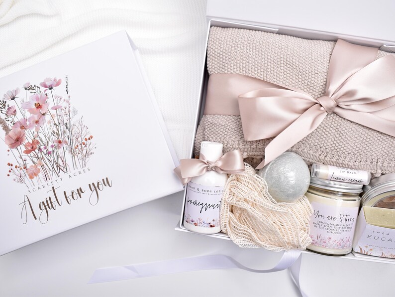 Op de afbeelding: Een witte geschenkdoos met een bloemenillustratie en de tekst Scarlett Acres A gift for you. Binnenin zit een beige gebreide deken met een satijnen lint, een flesje lotion, een bruisbal, lippenbalsem, een kaars en een netvormige spons.