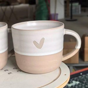 May include: A ceramic mug featuring a two-tone design with a matte beige lower section and a smooth white upper section. A small, simple heart shape is indented into the white portion of the mug. The mug includes a curved handle on the right side.
