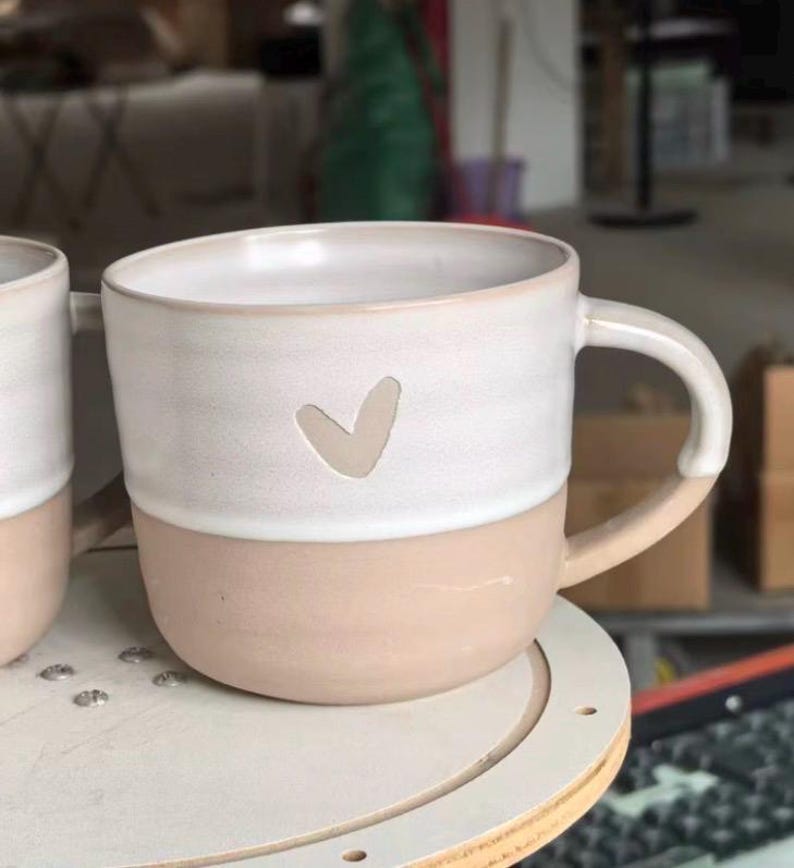 May include: A ceramic mug featuring a two-tone design with a matte beige lower section and a smooth white upper section. A small, simple heart shape is indented into the white portion of the mug. The mug includes a curved handle on the right side.