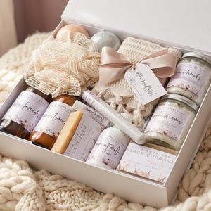 May include: A white gift box containing assorted bath and body products including glass jars with labels, a beige mesh sponge, a folded cream-colored textile with a pink ribbon and tag, a small rectangular soap bar, and two round bath bombs.