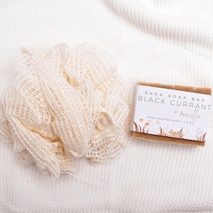 Op de afbeelding: Een beige mesh badspons naast een rechthoekig shea zeepblok met een wit label waarop Shea Soap Bar Black Currant + Vanilla Hand Crafted Soap 4.5 oz staat, voorzien van een botanische illustratie van bloemen en varens aan de onderzijde.