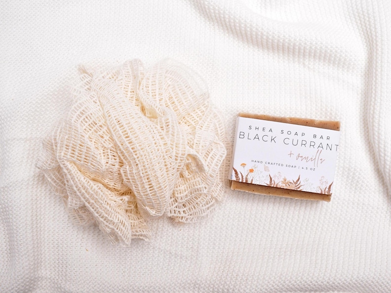 Op de afbeelding: Een beige mesh badspons naast een rechthoekig shea zeepblok met een wit label waarop Shea Soap Bar Black Currant + Vanilla Hand Crafted Soap 4.5 oz staat, voorzien van een botanische illustratie van bloemen en varens aan de onderzijde.