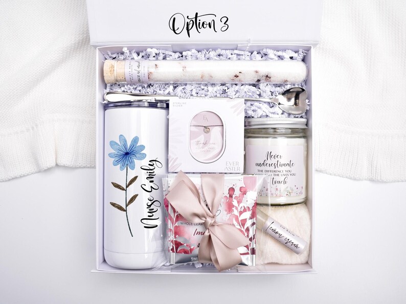 May include: A white gift box labeled Option 3 containing a white tumbler with a blue flower and the text Nurse Emily, a glass tube of bath salts, a silver spoon, a small jewelry box, a candle, a lip balm, and a gift bag with a pink ribbon.
