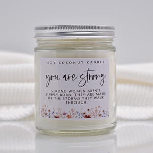 Op de afbeelding: Een kaars in een doorzichtige glazen pot met een zilveren deksel en witte was. Het etiket bevat de tekst: SOY COCONUT CANDLE, you are strong, STRONG WOMEN AREN'T SIMPLY BORN. THEY ARE MADE OF THE STORMS THEY WALK THROUGH, met een bloemenrand onderaan.
