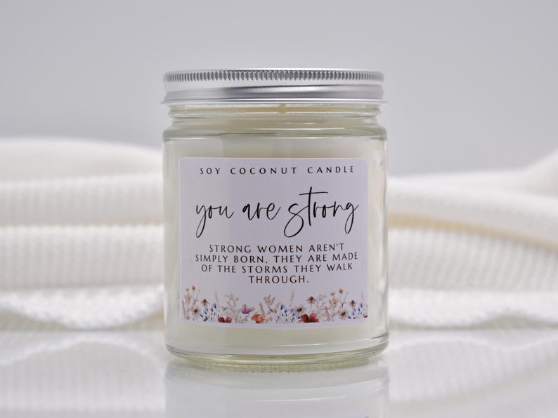 Op de afbeelding: Een kaars in een doorzichtige glazen pot met een zilveren deksel en witte was. Het etiket bevat de tekst: SOY COCONUT CANDLE, you are strong, STRONG WOMEN AREN'T SIMPLY BORN. THEY ARE MADE OF THE STORMS THEY WALK THROUGH, met een bloemenrand onderaan.
