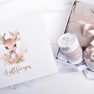 May include: A white gift box featuring a fawn illustration with pink flowers and the text A gift for you. Inside, a beige knit blanket, a white mug with fuzzy socks, a cylindrical container labeled Scarlett Acres Wellness, and a ribbon.