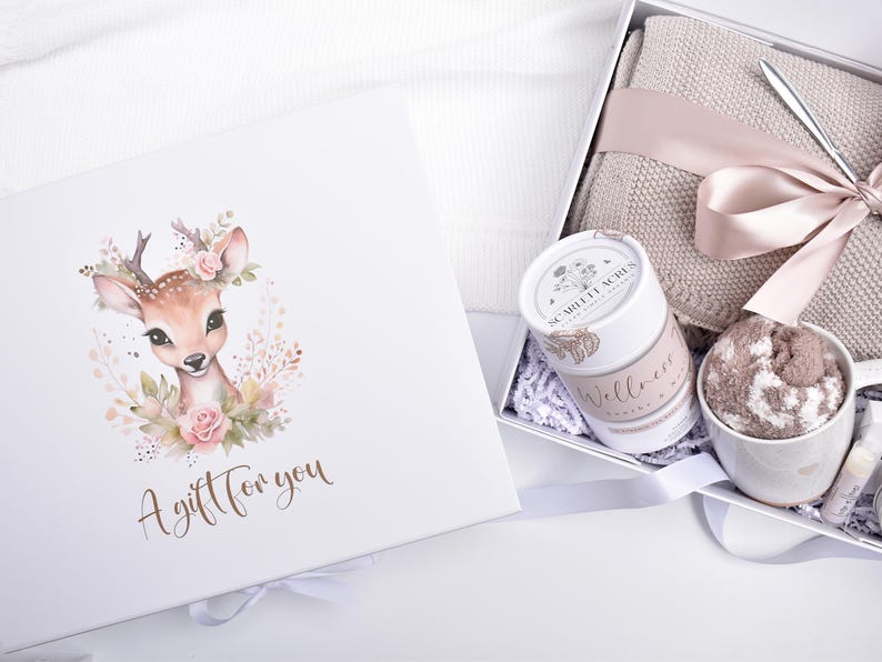 May include: A white gift box featuring a fawn illustration with pink flowers and the text A gift for you. Inside, a beige knit blanket, a white mug with fuzzy socks, a cylindrical container labeled Scarlett Acres Wellness, and a ribbon.