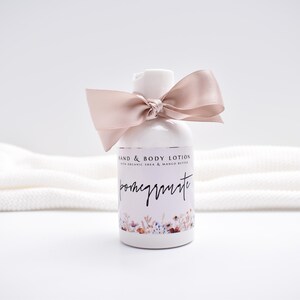 May include: A white plastic bottle of hand and body lotion with a dusty rose satin ribbon tied around the neck. The label features the text "HAND & BODY LOTION WITH ORGANIC SHEA & MANGO BUTTER" and "pomegranate" above a floral illustration.
