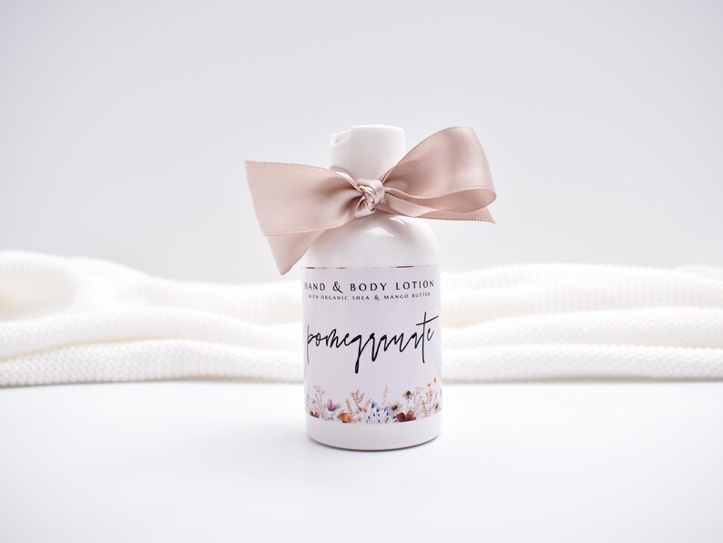May include: A white plastic bottle of hand and body lotion with a dusty rose satin ribbon tied around the neck. The label features the text "HAND & BODY LOTION WITH ORGANIC SHEA & MANGO BUTTER" and "pomegranate" above a floral illustration.