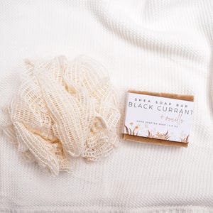 May include: A beige mesh bath sponge next to a rectangular shea soap bar with a white label that reads Shea Soap Bar Black Currant + Vanilla Hand Crafted Soap 4.5 oz, featuring a floral illustration at the bottom of the label.