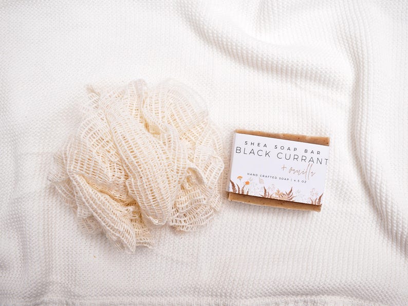 May include: A beige mesh bath sponge next to a rectangular shea soap bar with a white label that reads Shea Soap Bar Black Currant + Vanilla Hand Crafted Soap 4.5 oz, featuring a floral illustration at the bottom of the label.