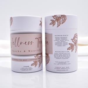 May include: Two cylindrical white cardboard tea canisters featuring copper-colored botanical illustrations. The front label reads Wellness Tea Soothe & Nourish, while the back lists ingredients including peppermint, spearmint, chamomile, and ginger.