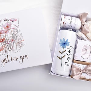 May include: A white gift box with a floral illustration and the text Scarlett Acres A gift for you. Inside is a white tumbler with a blue flower and the text Nurse Emily, a lotion bottle, lip balm, soap, a heart-shaped spoon, and a candle.