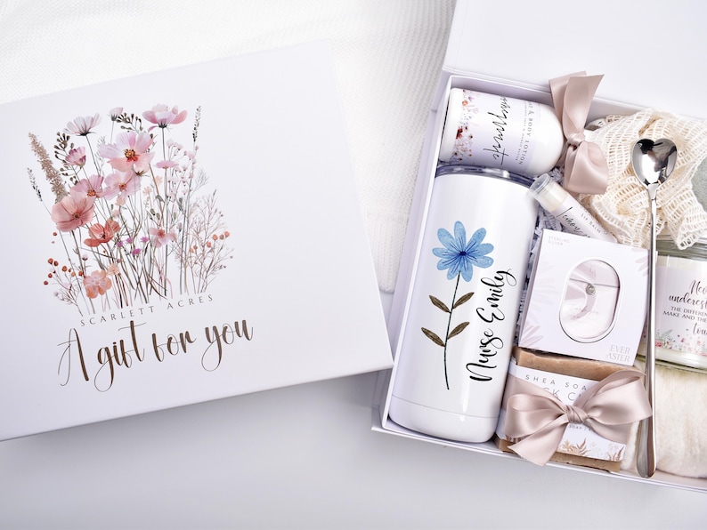 May include: A white gift box with a floral illustration and the text Scarlett Acres A gift for you. Inside is a white tumbler with a blue flower and the text Nurse Emily, a lotion bottle, lip balm, soap, a heart-shaped spoon, and a candle.