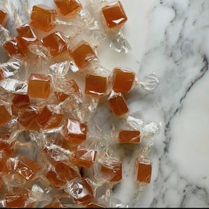 Op de afbeelding: Een verzameling vierkante, amberkleurige snoepjes, elk afzonderlijk verpakt in doorzichtig, gedraaid plastic folie. Elk stuk heeft een gladde, doorschijnende textuur en een consistente goudoranje tint, losjes verspreid over het beeldvlak.