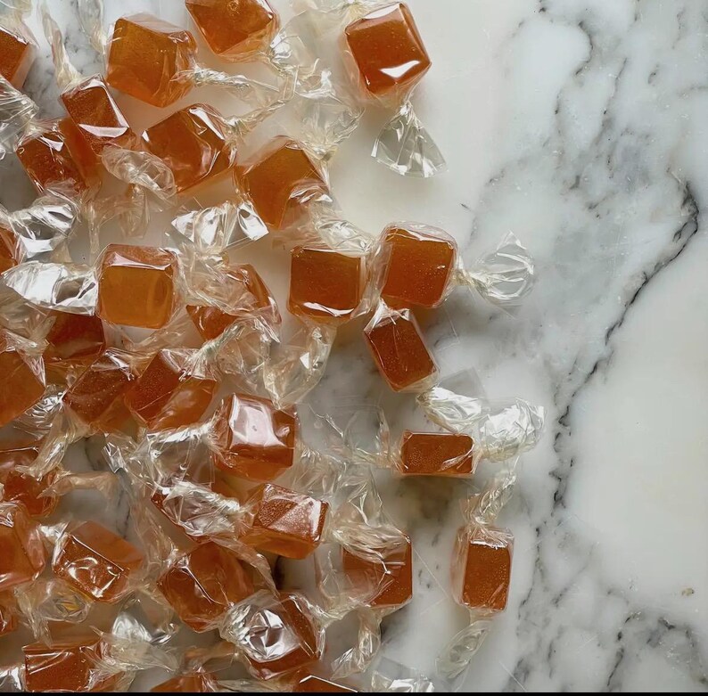 Op de afbeelding: Een verzameling vierkante, amberkleurige snoepjes, elk afzonderlijk verpakt in doorzichtig, gedraaid plastic folie. Elk stuk heeft een gladde, doorschijnende textuur en een consistente goudoranje tint, losjes verspreid over het beeldvlak.