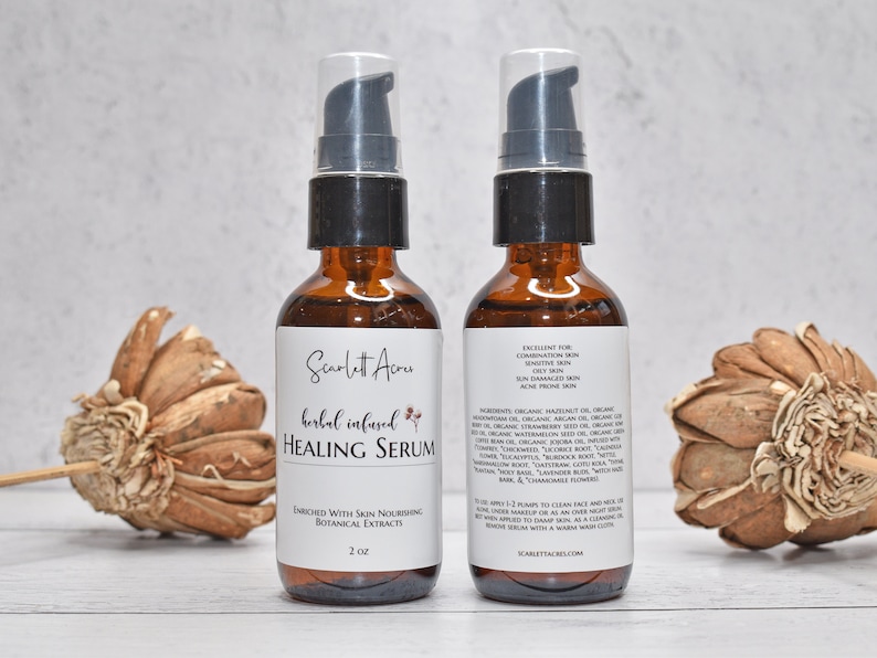 May include: Two amber glass bottles with black pump tops filled with a clear liquid. The bottles are labeled "Scarlett Acru Herbal Infused Healing Serum" and list ingredients and instructions for use. The bottles are sitting on a white surface with dried flowers on either side.