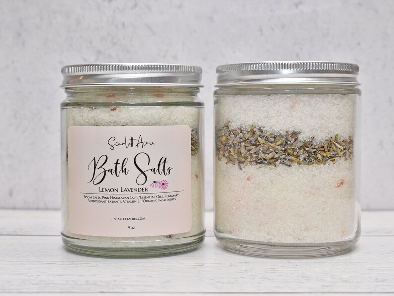 May include: Two glass jars with silver lids filled with bath salts. The jar on the left has a label that reads "Scarlett Acre Bath Salts Lemon Lavender Epsom Salt, Pink Himalayan Salt, Essential Oils, Rosemary Antioxidant Extract, Vitamin E, Organic Ingredients ScarlettAcres.com 9 oz."