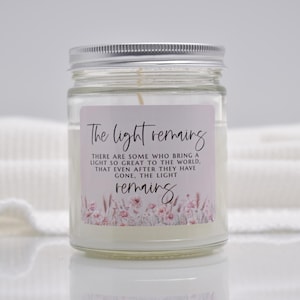 Puede incluir: Una vela en tarro de cristal transparente con tapa plateada. La vela tiene un interior de cera blanca y una etiqueta con el texto "The light remains". La etiqueta tambi&eacute;n presenta detalles florales en rosa y blanco.