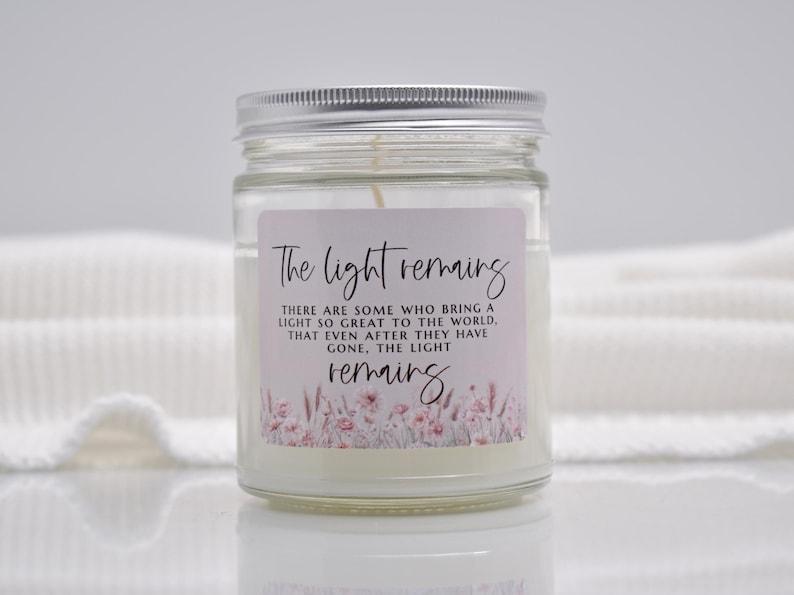 Puede incluir: Una vela en tarro de cristal transparente con tapa plateada. La vela tiene un interior de cera blanca y una etiqueta con el texto "The light remains". La etiqueta tambi&eacute;n presenta detalles florales en rosa y blanco.