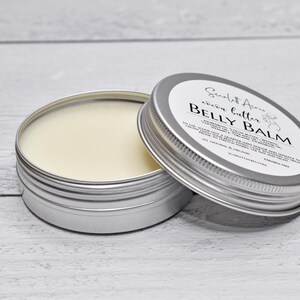 May include: A round silver metal tin containing off-white belly balm, with a matching lid resting beside it. The lid features a white label with black text reading Scarlett Acres, cocoa butter, BELLY BALM, ingredients, and usage instructions.