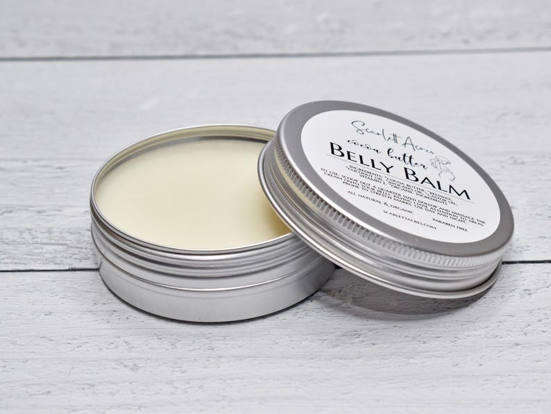 May include: A round silver metal tin containing off-white belly balm, with a matching lid resting beside it. The lid features a white label with black text reading Scarlett Acres, cocoa butter, BELLY BALM, ingredients, and usage instructions.