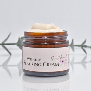 May include: A brown glass jar with a white label that reads "Wrinkle Repairing Cream" and "Scarlet Ann" in a pink floral design. The jar is filled with a white cream.